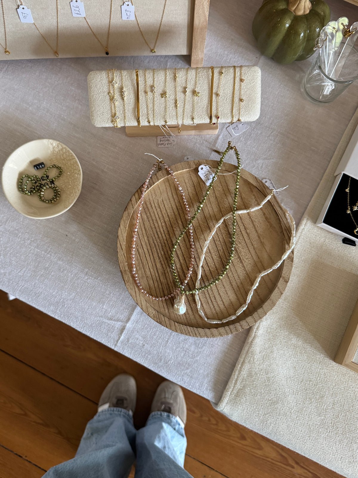 An overhead view of a jewelry display table with necklaces arranged on a round wooden tray, a small bowl, a cream fabric jewelry organizer, and a person's legs and shoes visible at the bottom. The setting includes a wood floor and a green decorative pumpkin.